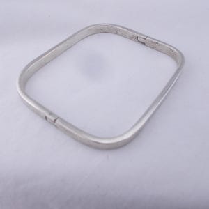 May include: A silver-coloured square bangle bracelet. The bracelet has a hinged clasp and a smooth, polished surface. The design is minimalist and modern, suitable for everyday wear. The bracelet's shape is a rounded square.