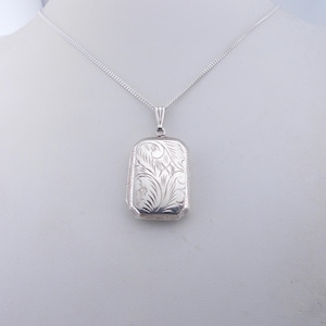 Könnte beinhalten: Eine silberne Halskette mit einem Anhänger, einer zarten Kette und einem rechteckigen Anhänger mit einem gravierten Blumenmuster.