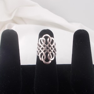 May include: A silver ring with an elaborate, interwoven pattern. The ring is shown on a black velvet finger stand against a white background. The ring's design consists of a series of looping, intertwined shapes, forming a complex pattern.
