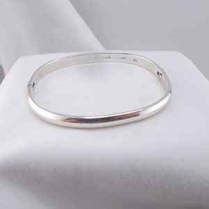 Può includere: Un bracciale rigido incernierato in metallo argentato lucido con un profilo liscio e arrotondato. L'interno della fascia presenta incisioni, tra cui il testo 925 che indica la composizione del metallo, e una piccola cerniera laterale.