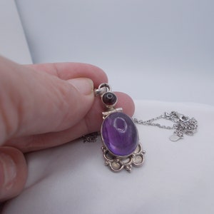 May include: A silver necklace with a purple amethyst and a dark red garnet pendant. The pendant has a detailed floral design.