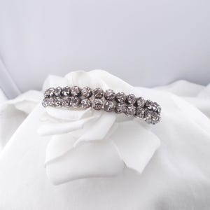 Könnte beinhalten: Ein silberfarbenes Armband mit zwei Reihen aus klaren, funkelnden Strasssteinen liegt auf einem weißen Stoff. Das Armband ist der Blickfang und zeigt sein aufwendiges Design und seine reflektierenden Eigenschaften. Der Hintergrund ist eine weiche, weiße und leicht strukturierte Oberfläche.