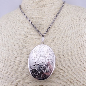 May include: Silver oval locket necklace with a detailed floral pattern. The locket is suspended from a silver chain and presented on a mannequin. The locket is approximately 5 cm in length.