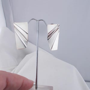 Puede incluir: Un par de pendientes de plata con un diseño geométrico. Los pendientes tienen forma de rectángulo con un borde curvo. Se muestran en un soporte.