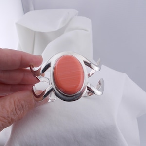 Può includere: Un bracciale a polsino in argento con una pietra ovale color corallo. Il bracciale presenta un design traforato decorativo con elementi curvi a forma di onda. Sfondo bianco.