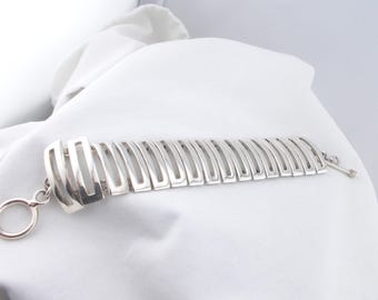 May include: A silver bracelet with a geometric design. The bracelet is made of multiple connected rectangular pieces, each with a cut-out in the centre. The bracelet has a simple, modern look.
