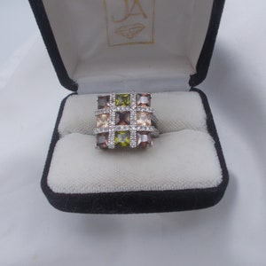 Puede incluir: Un anillo de forma cuadrada con una banda de plata y múltiples piedras preciosas cuadradas en tonos de marrón, verde y transparente. El anillo está en una caja de anillo de terciopelo negro.