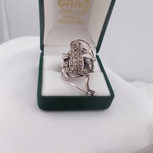 May include: A silver frog ring with red gemstone eyes and marcasite detailing. The ring is displayed in a green velvet jewelry box. The frog is the focal point, with its legs extended and body covered in small stones.