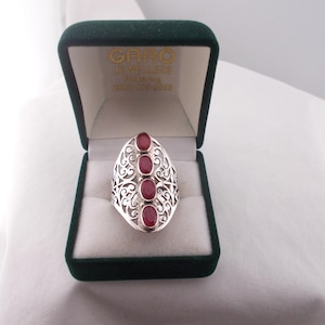 May include: A silver ring with an oval filigree design, set with four oval, deep red gemstones. The ring is displayed in a dark green jewellery box with the text "CARO JEWELLERS" visible on the inside of the lid.