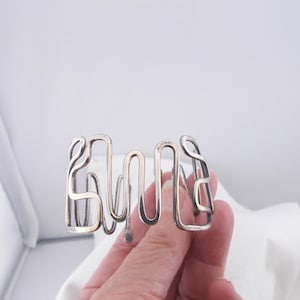 May include: A silver bracelet with a unique, abstract design. The bracelet is made from a single piece of metal that is bent and twisted into a series of loops and curves.