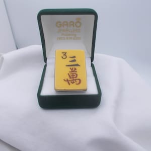 Może przedstawiać: Żółto-czerwona kostka mahjongowa z numerem 3 i chińskimi znakami w zielonym aksamitnym pudełku z napisem "GARO JEWELLERS" i numerem telefonu.