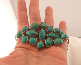 Op de afbeelding: Een zilveren armband met meerdere turkooizen stenen in verschillende vormen, waaronder ovalen, vierkanten en driehoeken. De stenen zijn gezet in zilveren zettingen en verbonden door een zilveren ketting.