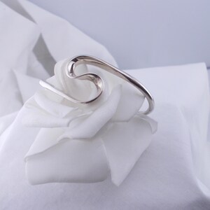 Peut inclure: Un bracelet argenté au design abstrait et fluide. Le bracelet est incurvé et repose sur un tissu blanc, créant un contraste entre le métal et le fond doux. Le design du bracelet rappelle une vague ou une lettre stylisée.