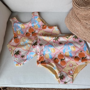 May include: A two-piece swimsuit with a patchwork design featuring strawberries, oranges, and bows. The top is a halter style and the bottoms are high-waisted.