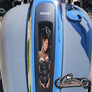 Könnte beinhalten: Ein Motorrad-Benzintank mit einem schwarzen und silbernen Road Glide-Emblem. Eine Schwarzweißillustration einer Frau in einem schwarzen Korsett und einem Hut befindet sich auf dem Tank.