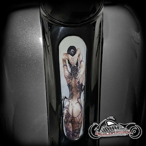 Peut inclure: Un réservoir d'essence de moto noir avec un autocollant décoratif représentant une femme vêtue d'une robe à motifs noirs et blancs. L'autocollant est sur le côté du réservoir et est entouré d'une bordure noire. La moto est également décorée des mots "American Freedom" en lettres blanches.