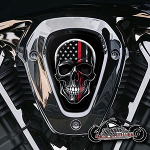 Könnte beinhalten: Ein schwarz-weißer Schädel mit einem roten Streifen in der Mitte, überlagert von einer amerikanischen Flagge. Der Schädel befindet sich auf einem schwarzen Motorradeinlassdeckel.