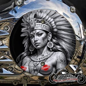 May include: A chrome motorcycle component with a monochrome illustration of a woman adorned with a feathered headdress and tribal markings. The image is set within a polished metal frame, with the text "AMERICAN FREEDOM".