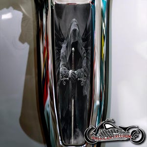 May include: A black and white image of a hooded figure with wings holding a sword. The figure is standing in front of a dark background. The image is in a chrome frame.