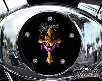 Op de afbeelding: Een zwart motoraccessoire met een gouden kruis en een ontwerp met gevouwen handen. Het accessoire heeft het woord "Blessed" in blauwe en witte letters.