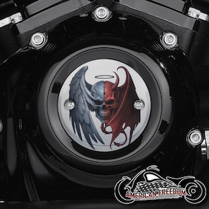 Puede incluir: Una cubierta de motor de motocicleta con un gráfico detallado de una calavera, dividida en mitades de ángel y diablo. El lado del ángel presenta un halo y un ala, mientras que el lado del diablo tiene cuernos y un ala. La cubierta está sobre un fondo negro.