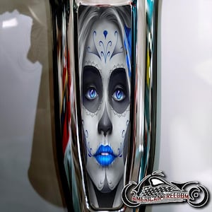 Puede incluir: Primer plano de un tanque de gasolina de motocicleta con una pegatina decorativa que presenta el rostro de una mujer con ojos azules y un diseño de maquillaje de calavera de azúcar. La pegatina tiene un patrón floral blanco y azul.