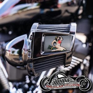 May include: A chrome motorcycle part with a rectangular inset featuring a pin-up girl illustration. The illustration depicts a woman in a red top, seated on a bomb. The text "CENSORED" is visible. The motorcycle part is mounted on a black motorcycle.