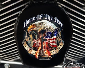 May include: A black motorcycle gas tank cover featuring a graphic of a bald eagle, an American flag, and a soldier's boot. The text "Home of the Free Because of the Brave" is written in a banner around the graphic.