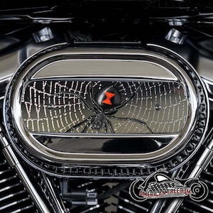 Pode incluir: Close-up de uma aranha preta com um relógio de areia vermelho nas costas, sentada em uma teia em uma tampa de filtro de ar de motocicleta cromada. A tampa é redonda e tem um acabamento brilhante.