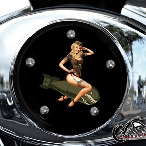 May include: A chrome motorcycle gas cap cover with a black background featuring a pin-up girl in a military uniform riding a cartoon bomb with a shark fin. The text "AMERICAN FREEDOM" is visible at the bottom of the image.