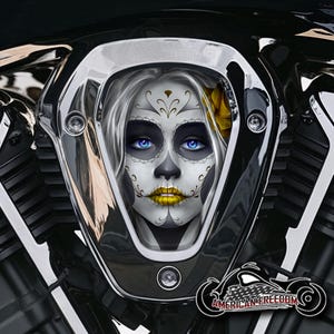 Könnte beinhalten: Ein verchromter Motorradmotordeckel mit einem stilisierten Gesicht im Sugar-Skull-Stil. Das Gesicht hat blaue Augen und goldene Lippen. Eine gelbe Blume ist zu sehen. Das Logo unten lautet "AMERICAN FREEDOM".