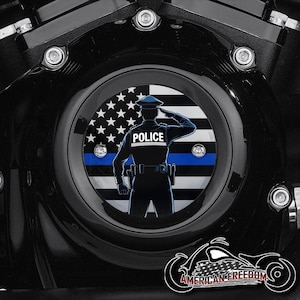 Puede incluir: Tapa de motor de motocicleta negra con un emblema circular que presenta a un oficial de policía saludando frente a un diseño de bandera estadounidense. La palabra "POLICE" es visible en el uniforme del oficial. El emblema está rodeado por una carcasa de motor negra.