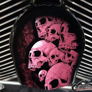 May include: A black motorcycle gas tank lid with a pink skull design. The skulls are layered on top of each other and have a distressed look.
