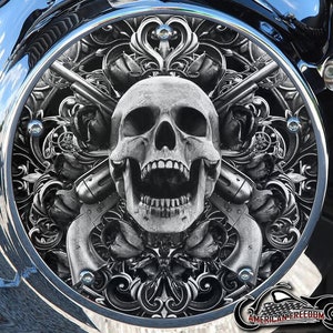 May include: A black and white skull with crossed pistols and ornate floral designs. The skull is grinning with its mouth open and teeth showing. The design is on a round, silver-colored background.