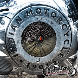 Pode incluir: Close-up de uma tampa de motor de motocicleta cromada com um design de teia de aranha preta e uma aranha preta no centro. A tampa tem as palavras "INDIAN MOTORCYCLE" e "1901" inscritas nela.