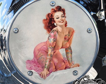 May include: A round, chrome motorcycle cover with a pin-up girl image. The girl is wearing a pink dress and has red hair and tattoos on her arms. The cover has six chrome screws around the edge.