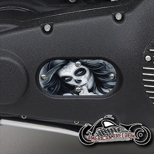 May include: A black motorcycle part with a decorative cover featuring a black and white illustration of a woman with a sugar skull makeup design. The woman is wearing a black top and has long, flowing hair.