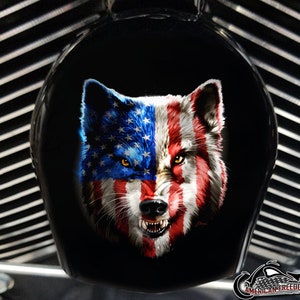 May include: A black motorcycle part with a detailed graphic of a wolf's face. The wolf's fur is painted with the American flag design, featuring red and white stripes on one side and blue with white stars on the other. The wolf has yellow eyes and bared teeth.