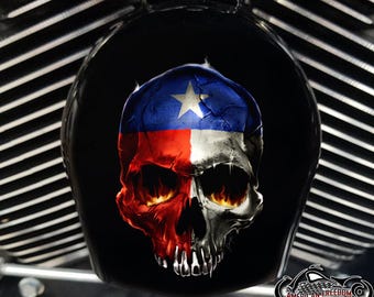 May include: Black motorcycle component featuring a skull design. The skull is painted in the colours of the Texas flag: blue, white, and red. Flames emerge from the eye sockets. The logo "American Freedom" is at the bottom.
