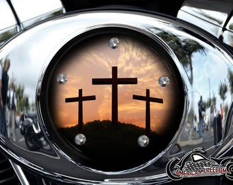 May include: A chrome motorcycle gas cap with a black insert featuring three crosses silhouetted against a sunset sky. The crosses are centered in the image and are the focal point. The gas cap is round and has a reflective surface.