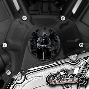 May include: A black motorcycle engine cover with a circular cutout featuring a black and white image of a hooded figure with wings holding a sword.