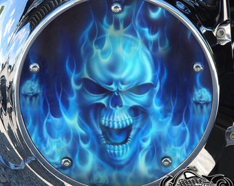 May include: A round, chrome motorcycle cover with a blue and white skull and flames design. The skull is outlined in black and has a white mouth with teeth. The flames are blue and white and are surrounding the skull.