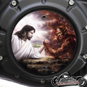 Puede incluir: Una cubierta redonda de motocicleta negra con una imagen colorida de Jesús y el Diablo luchando en un pulso. La imagen está sobre un fondo de cielo tormentoso y un volcán en erupción.