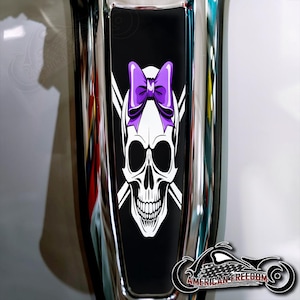 May include: A black and white skull graphic with a purple bow, set against a black background. The design includes crossed bones and is displayed on a chrome surface. The image also features the text "AMERICAN FREEDOM".