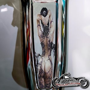 May include: A chrome motorcycle gas tank with a decorative sticker of a woman in a black net dress. The sticker is a detailed illustration with a silver background.