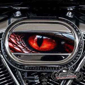 May include: Chrome motorcycle air cleaner cover featuring a vibrant red and black reptilian eye design. The eye is set against a dark background, with the words "AMERICAN FREEDOM" and a motorcycle graphic at the bottom.