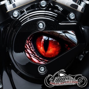 Può includere: Coperchio motore per moto nero con una grafica rossa e nera di un occhio di rettile. L'occhio è dettagliato con squame e una superficie riflettente. Il coperchio è fissato con bulloni argentati e presenta il testo "AMERICAN FREEDOM".