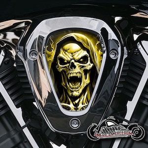 Könnte beinhalten: Ein verchromter Motorradeinlassdeckel mit einer gelben Totenkopfgrafik. Der Totenkopf trägt eine Kapuze und hat einen bedrohlichen Ausdruck. Die Grafik ist auf einem schwarzen Hintergrund.