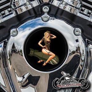 May include: A chrome motorcycle engine cover with a round, black insert featuring a pin-up girl in a military uniform sitting on a bomb with a shark mouth. The text "AMERICAN FREEDOM" is visible at the bottom of the image.