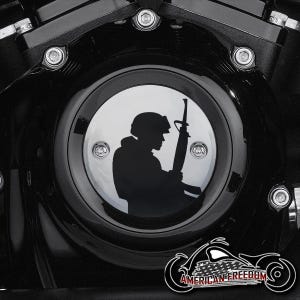 Puede incluir: Cubierta negra del motor de una motocicleta con un emblema plateado que representa la silueta de un soldado sosteniendo un rifle. El emblema está rodeado por un marco circular negro con pernos plateados. Las palabras "AMERICAN FREEDOM" se muestran debajo.